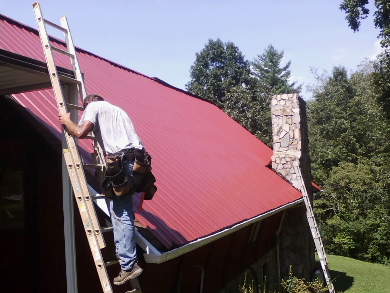 Roofer on a ladder beside a red metal roof and stone chimney — Soffit & Fascia Repair by Roofing Brothers in Francis Creek